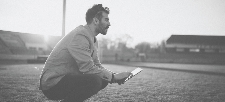 A man holding an ipad crouches on a sports field, looking pensively out of frame